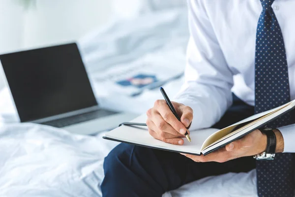 Vista cortada de empresário escrevendo em diário enquanto sentado na cama com laptop — Fotografia de Stock