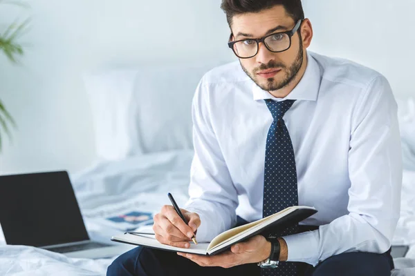 Bel homme d'affaires écrit dans un journal tout en étant assis sur le lit avec ordinateur portable — Photo de stock