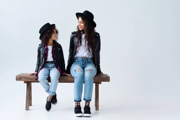 Happy mother and daughter in similar clothes sitting on bench together isolated on grey — Stock Photo