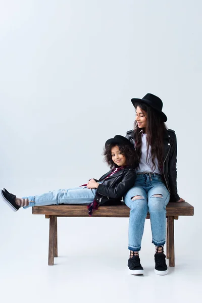 Mãe e filha em roupas semelhantes sentadas no banco juntas isoladas em cinza — Fotografia de Stock