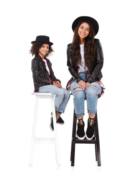 Stylish mother and daughter in hats and leather jackets sitting on chairs isolated on white — Stock Photo
