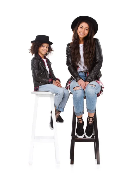 Elegante madre e hija en sombreros y chaquetas de cuero sentados en sillas aisladas en blanco - foto de stock