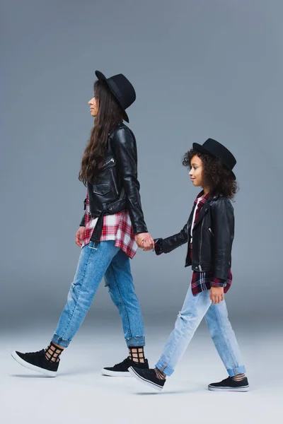 Vista lateral de la madre y la hija con estilo caminando juntas y tomados de la mano en gris - foto de stock