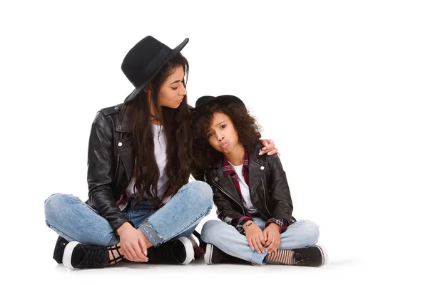 Beautiful mother and daughter in leather jackets and hats sitting on floor isolated on white — Stock Photo
