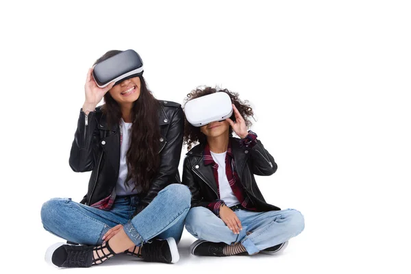 Excited young mother and daughter in virtual reality headsets sitting on floor isolated on white — Stock Photo