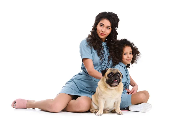 Hermosa joven madre e hija sentado en el suelo con pug aislado en blanco - foto de stock