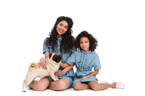 Madre e hija sentadas en el suelo con pug aislado en blanco - foto de stock