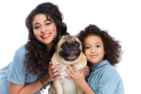 Madre e hija abrazando adorable pug y mirando a la cámara aislada en blanco - foto de stock