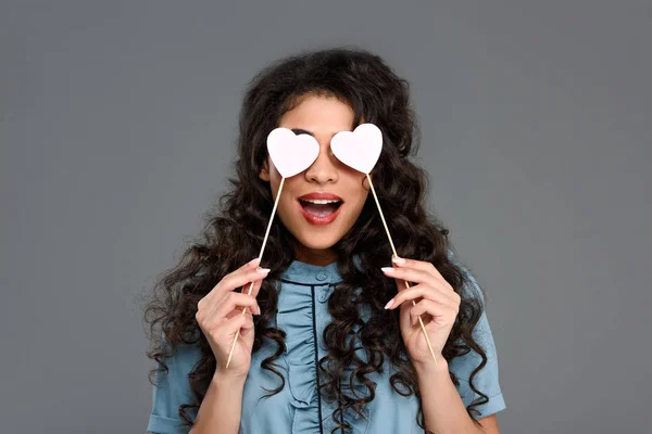 Excited young woman covering eyes with hearts on sticks isolated on grey — Stock Photo