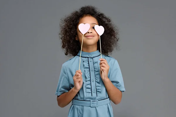 Niño feliz cubriendo los ojos con corazones signos aislados en gris - foto de stock