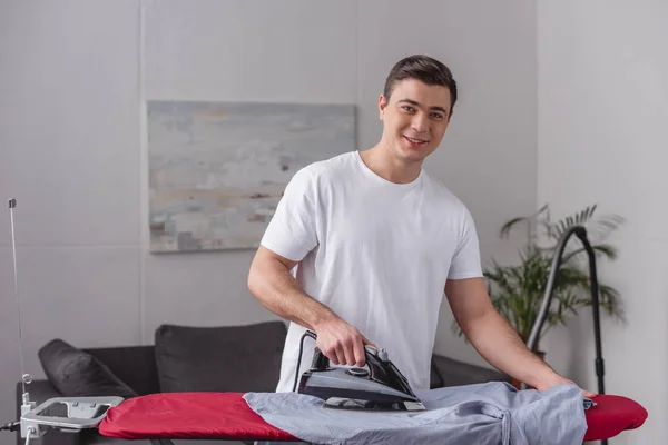 Souriant bel homme repassage chemise sur planche à repasser dans le salon — Photo de stock