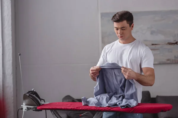 Homem bonito segurando camisa para passar na sala de estar — Fotografia de Stock