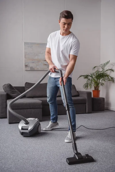 Hombre guapo alfombra de limpieza en la sala de estar con aspiradora - foto de stock