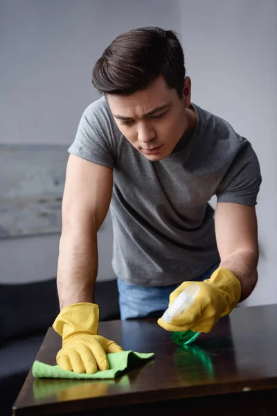 Handsome man cleaning table in living room with spray bottle and rag — Stock Photo