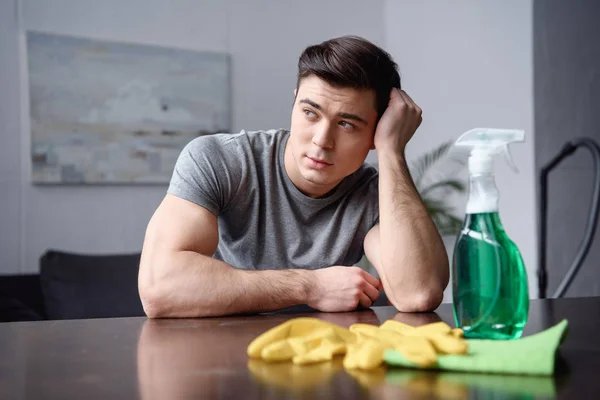 Hombre guapo sentado en la mesa con cosas de limpieza y mirando hacia otro lado en la sala de estar - foto de stock