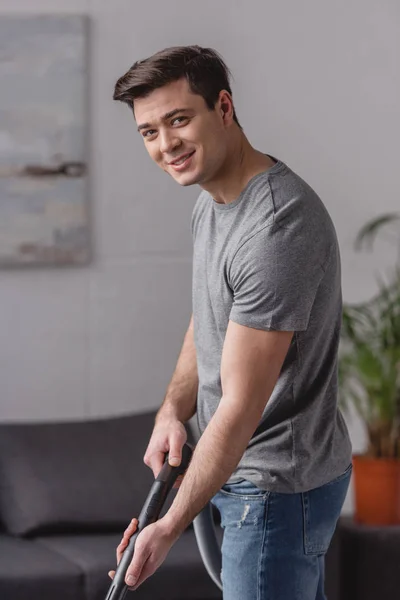 Handsome man cleaning carpet with vacuum cleaner and looking at camera in living room — Stock Photo