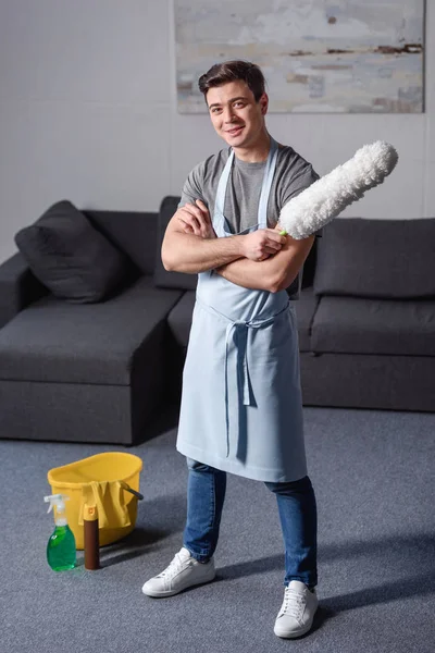 Усміхнений красивий чоловік стоїть і тримає щітку для чищення пилу у вітальні — стокове фото