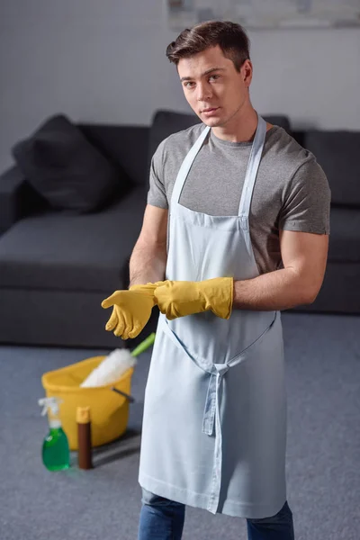 Hombre guapo con guantes de goma para limpiar y mirar a la cámara en la sala de estar - foto de stock