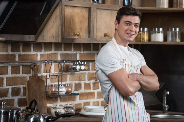 Sonriente hombre guapo de pie con los brazos cruzados en la cocina - foto de stock