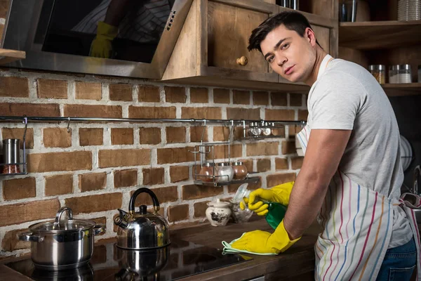 Bel homme nettoyage cuisine avec vaporisateur et chiffon et regarder la caméra — Photo de stock