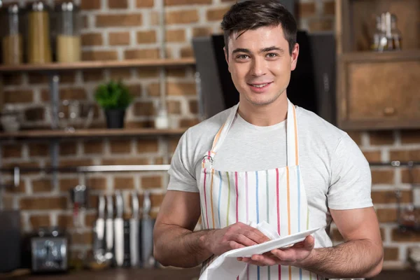 Hombre guapo limpiando plato con toalla y mirando a la cámara en la cocina — Stock Photo