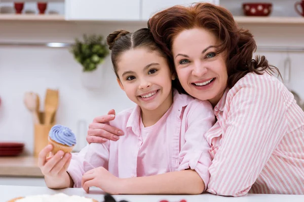 Abuela y pequeña nieta con cupcake sentado en la cocina y mirando a la cámara - foto de stock