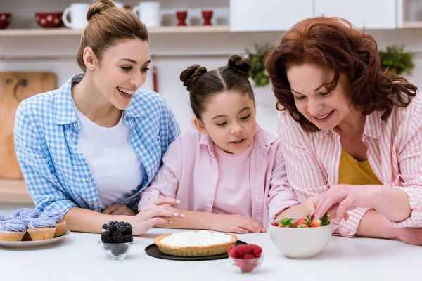 Three generations of women decorating dessert with berries together at kitchen — Stock Photo