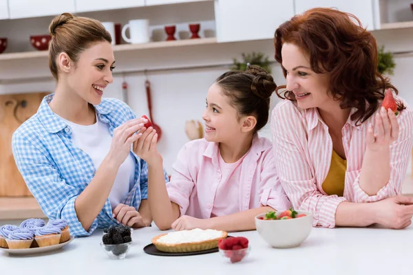 Tres generaciones de mujeres riendo decorando el postre con bayas juntas en la cocina - foto de stock