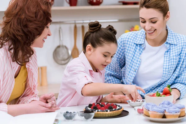 Drei Generationen schöner Frauen dekorieren gemeinsam in der Küche ein Dessert mit Beeren — Stockfoto