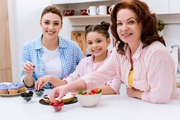 Trois générations de femmes décorant le dessert avec des baies ensemble à la cuisine et regardant la caméra — Photo de stock
