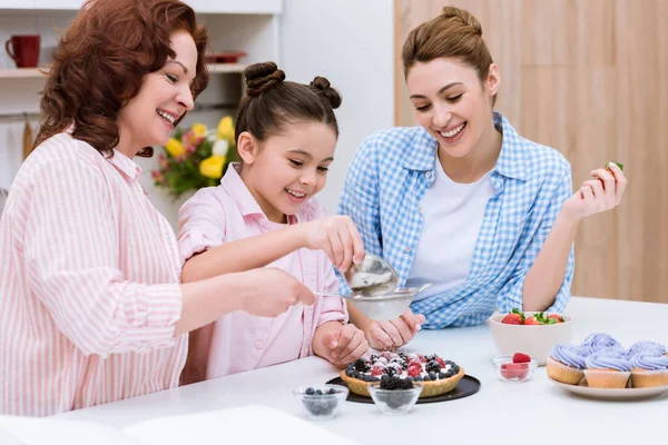 Tres generaciones de mujeres vertiendo azúcar en tarta con bayas juntas en la cocina - foto de stock