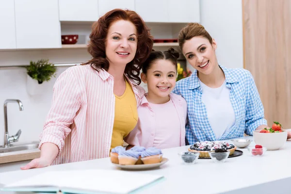 Tres generaciones de mujeres con postres en la cocina mirando la cámara - foto de stock