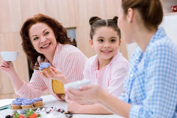 Tres generaciones de mujeres pasando tiempo juntas en la cocina y comiendo postres con té en la cocina - foto de stock