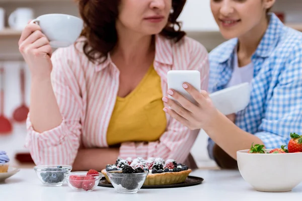 Cropped shot of woman and her mother drinking coffee with sweets and using smartphone together — Stock Photo