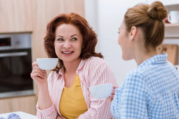 Young woman and her mother drinking coffee at kitchen and chatting — Stock Photo