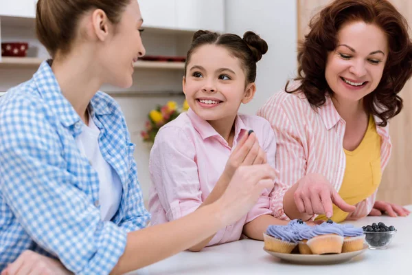 Three generations of women decorating cupcakes with blueberries together at kitchen — Stock Photo