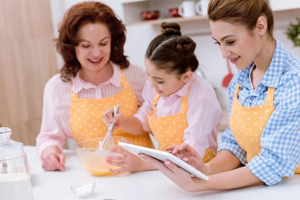 Großmutter und Enkelin kochen zusammen, während die Mutter in der Küche ein Tablet benutzt — Stockfoto