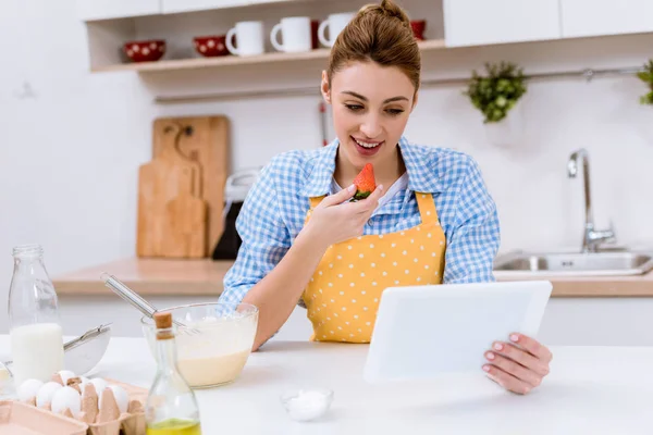 Attractive young woman eating strawberry and using tablet at kitchen while cooking — Stock Photo