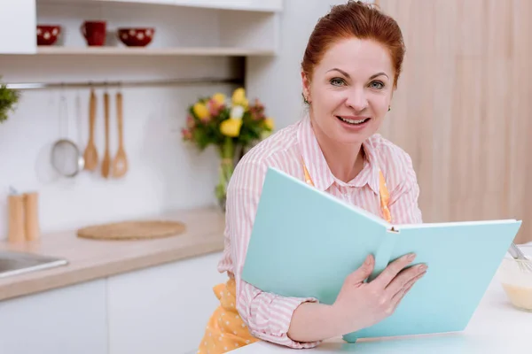 Щаслива зріла жінка в фартусі з книгою рецептів — стокове фото