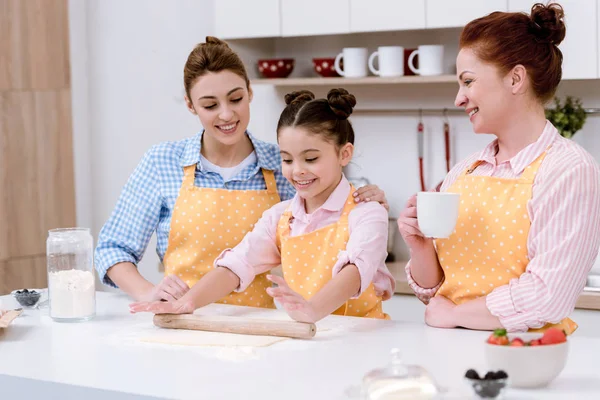 Tres generaciones de mujeres felices rodando masa para galletas juntos en la cocina - foto de stock