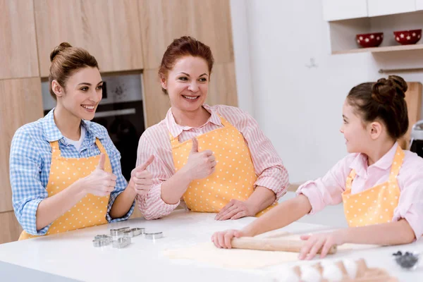 Grandmother and mother showing thumbs up to daughter while she preparing dough for cookies at kitchen — Stock Photo