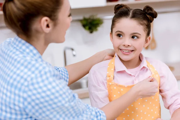 Mãe vestindo sua filha em avental na cozinha — Fotografia de Stock