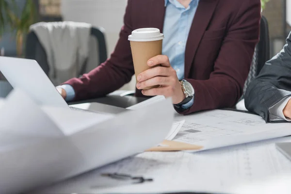 Recortado tiro de arquitecto con taza de papel de café trabajando con planos de construcción - foto de stock