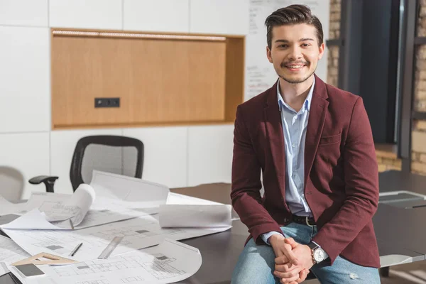 Giovane architetto sorridente seduto sul tavolo da lavoro con piani di costruzione in ufficio — Foto stock