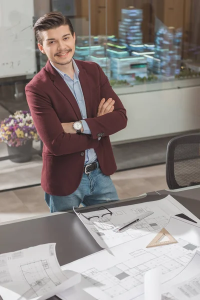 Sonriente joven arquitecto con brazos cruzados cerca del lugar de trabajo en la oficina - foto de stock