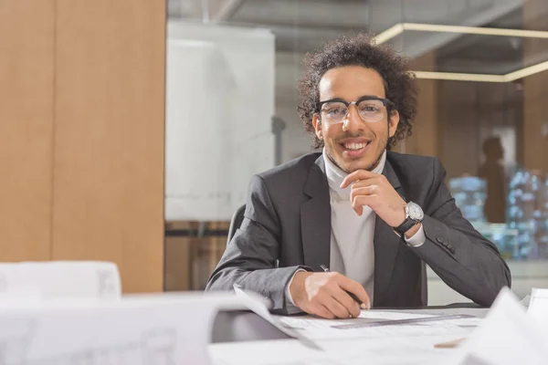 Sonriente joven arquitecto con planos de construcción sentado en el lugar de trabajo en la oficina - foto de stock