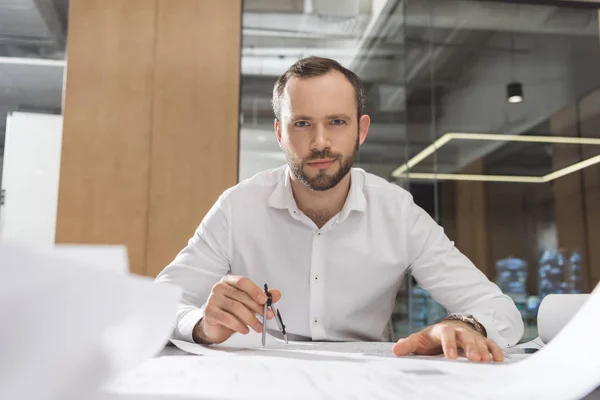 Bonito arquiteto bem sucedido com bússola trabalhando em planos de construção no escritório — Fotografia de Stock