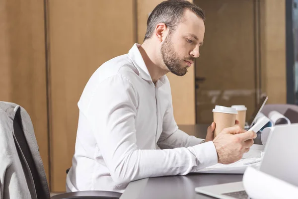 Serious architect with paper cup of coffee using smartphone at office — Stock Photo