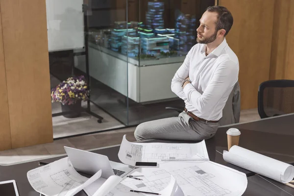 Arquitecto guapo sentado en la mesa con los brazos cruzados en la oficina - foto de stock