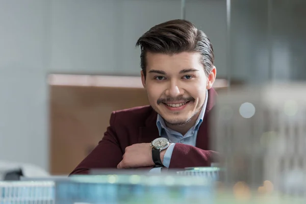 Bonito sorrindo arquiteto olhando para câmera no escritório atrás de modelos de construção — Fotografia de Stock